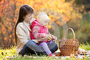 Kid and mother sit with apples basket outdoors in autumnal park